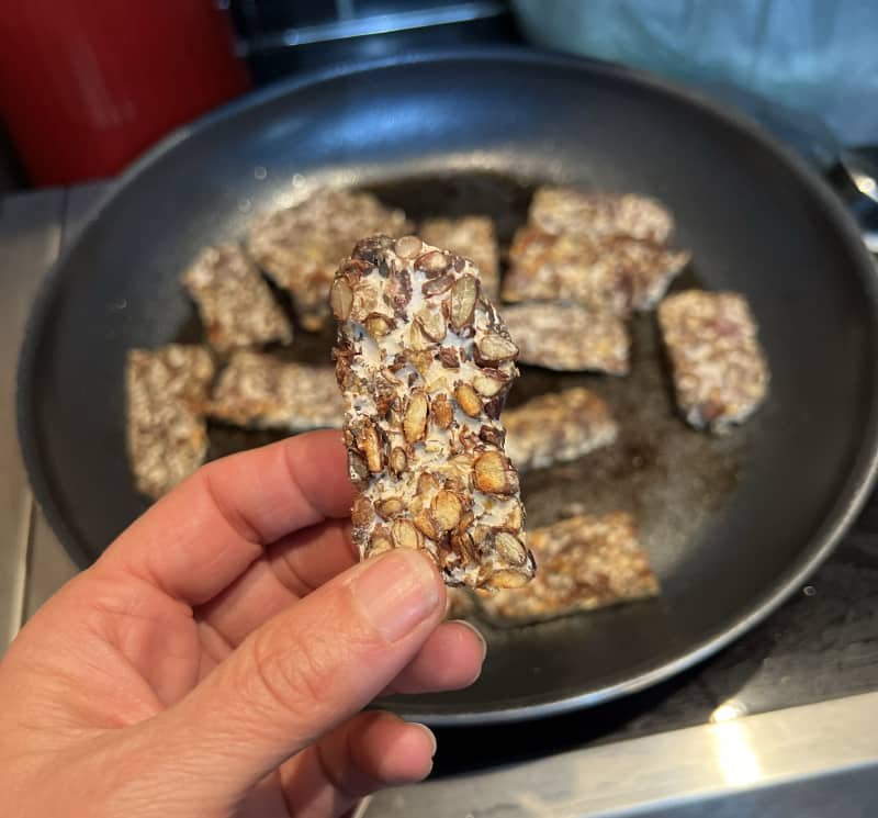 a hand showing a piece of fresh tempeh that has just been cooked with more tempeh pictured in the frying pan behind it.
