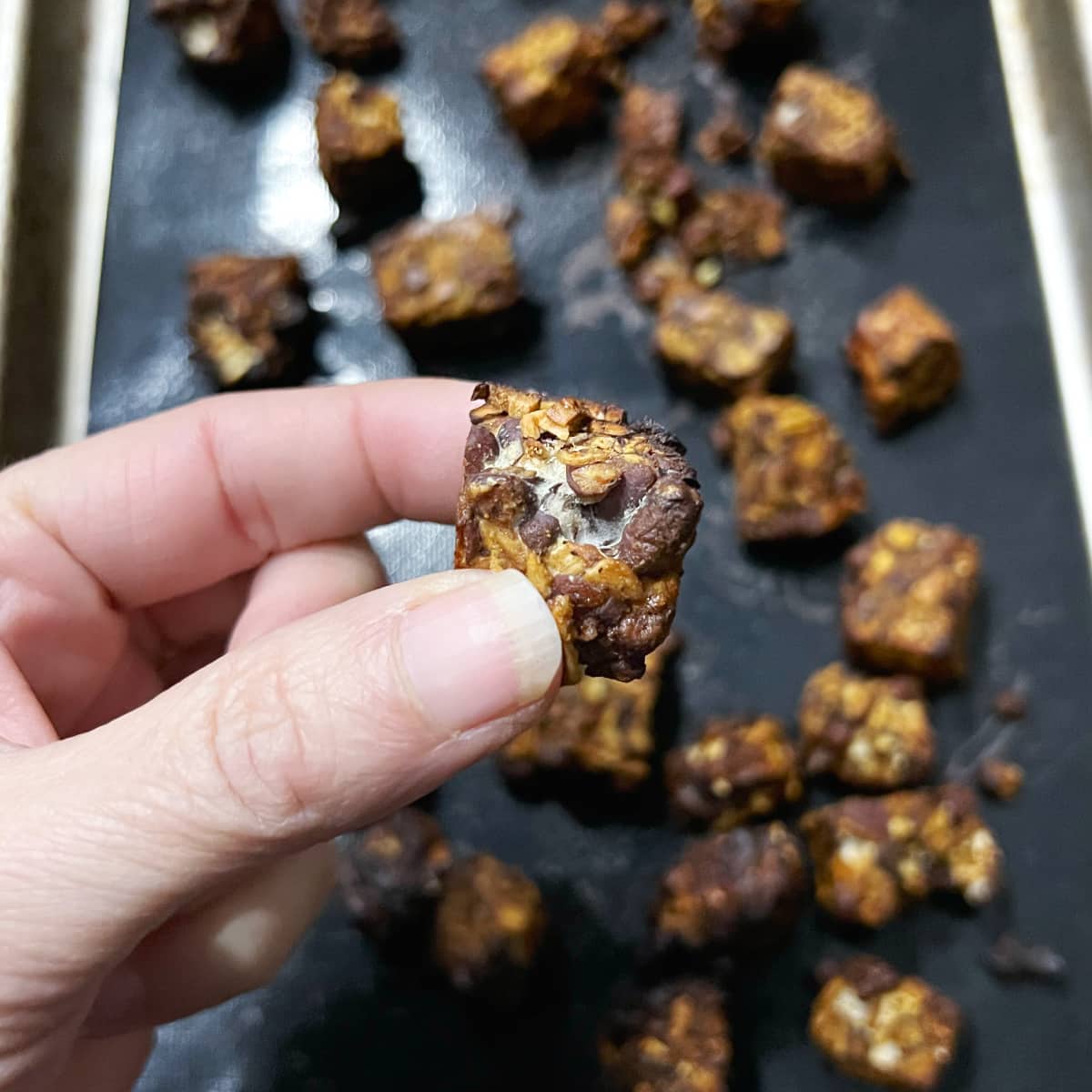 a hand holding a piece of balsamic dijon glazed tempeh with a baking sheet full of them in the background.