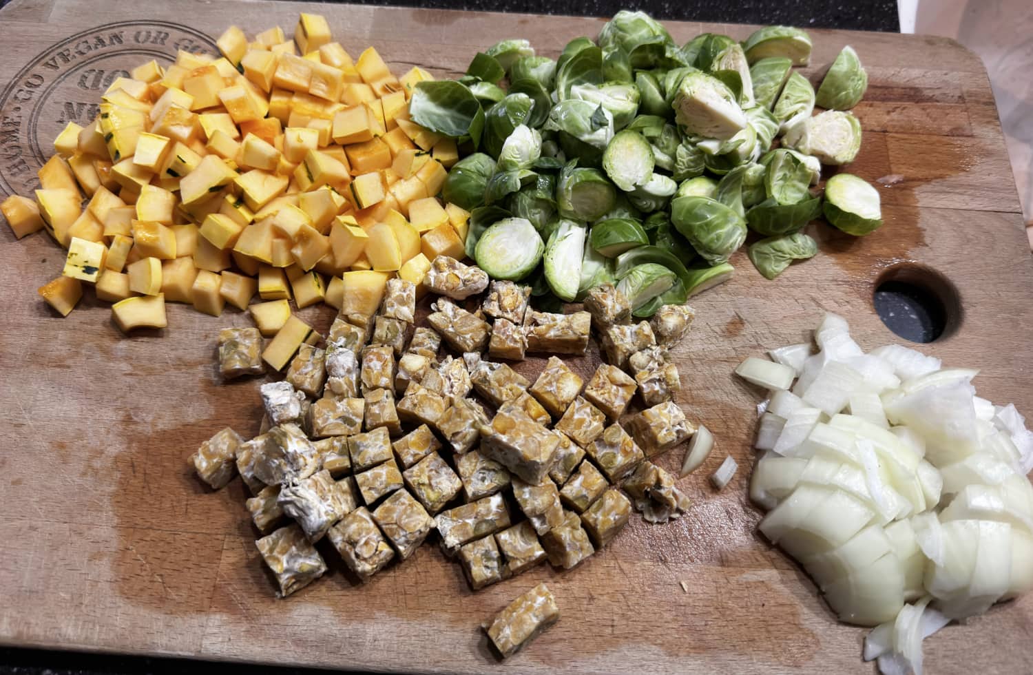 prepped delicata squash, brussels sprouts, tempeh and onions on a wood cutting board.
