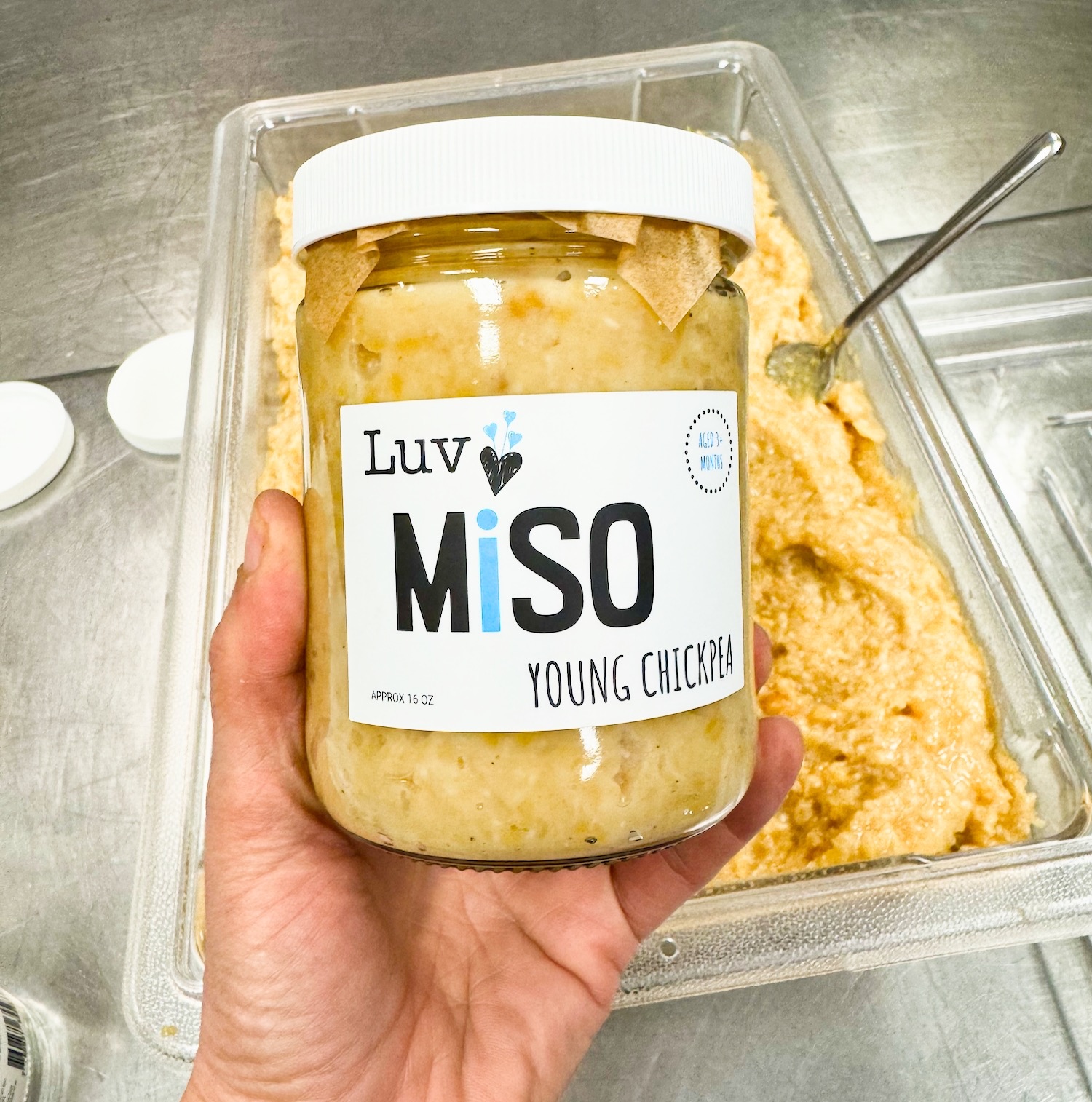 packaging-chickpea-miso hand holding a jar of Luv chickpea miso with a container of the miso in the background.